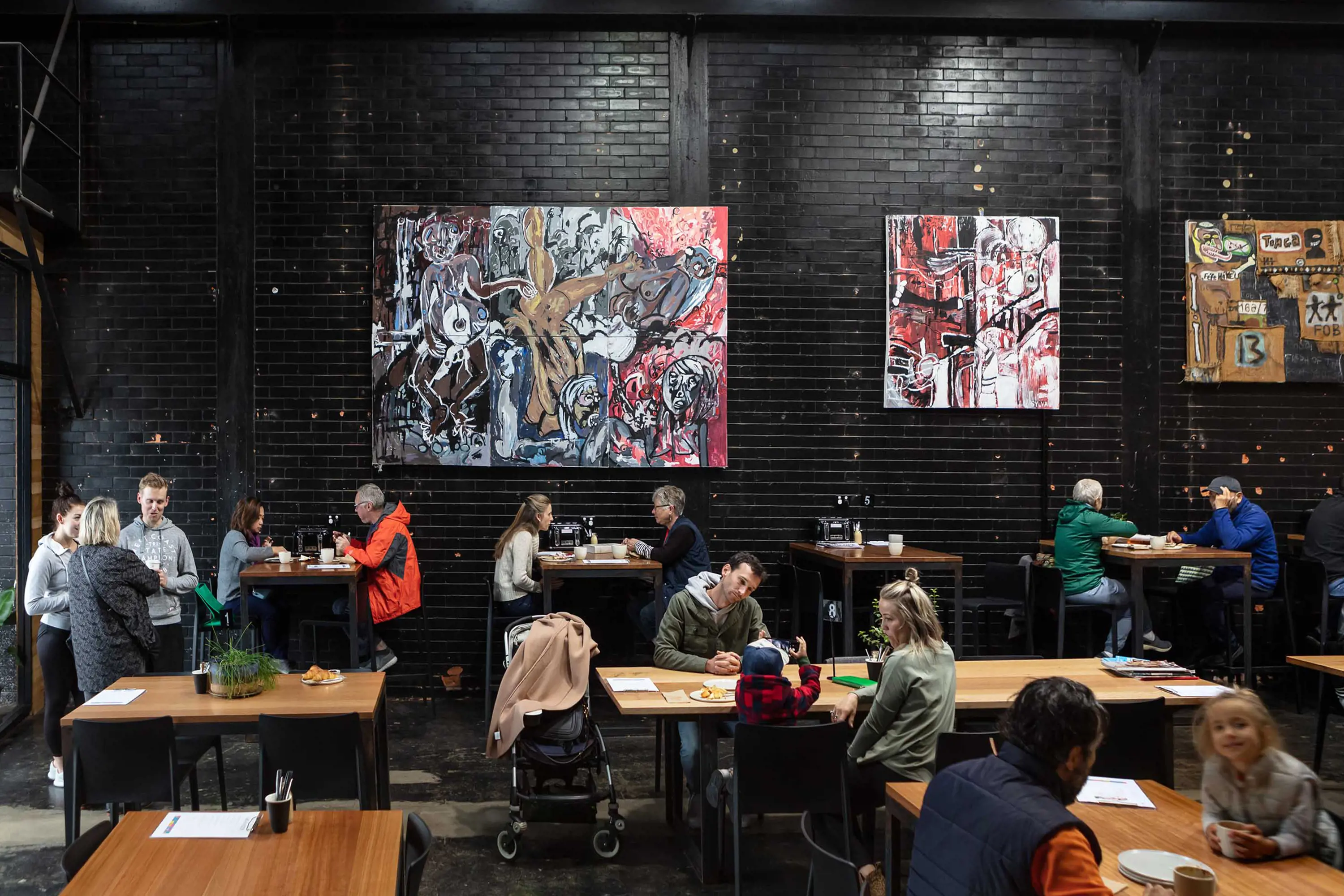 Groups of people sit at tables in a cafe within a converted factory space. Contemporary artwork is hung form the brick walls, which have been painted black.