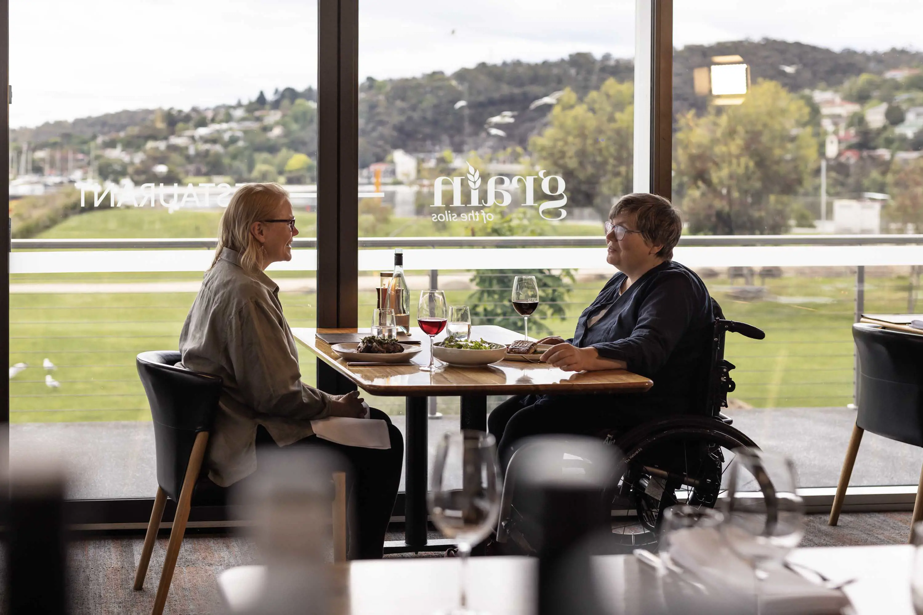 Two people sit at a table, one in a wheelchair, in the tall windows of a restaurant overlooking nearby parkland and a river.