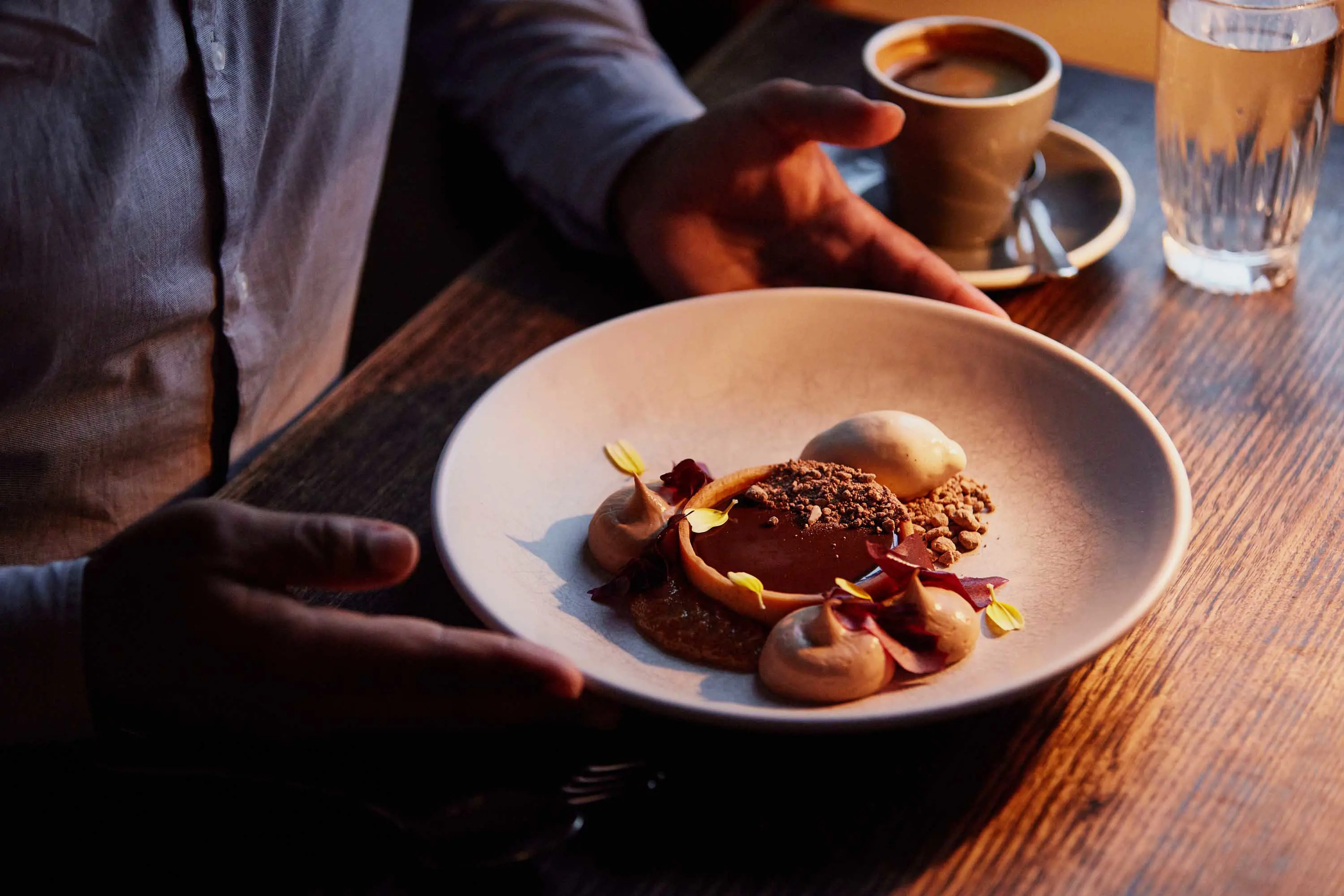 A waiter places a earthenware dish with a dessert onto a dark wooden table.