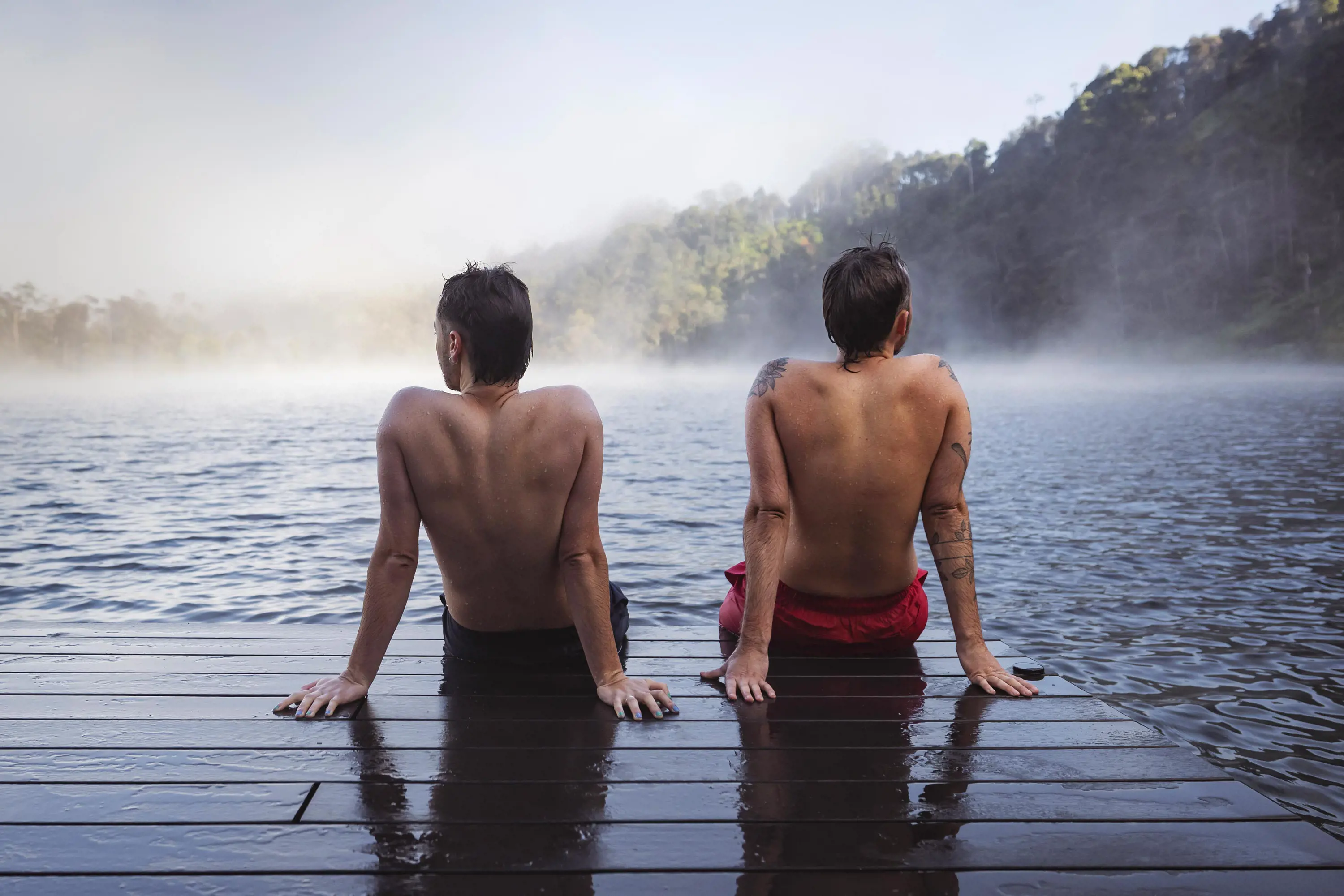 Two young men sit on the end of wooden decking looking out over a misty lake. 