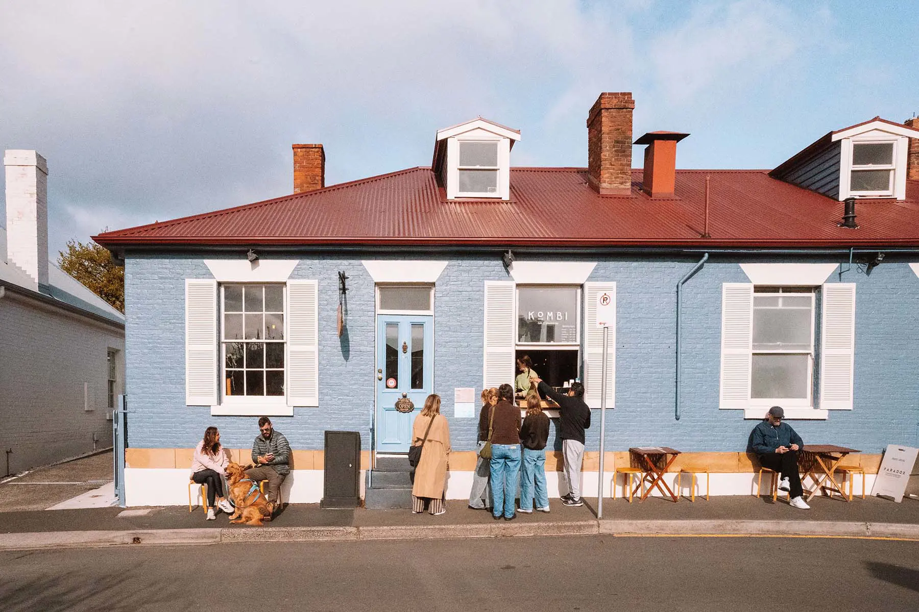 A cafe in a little blue painted brick building with white shutters. People sit at tables on the footpath and order through an open window.