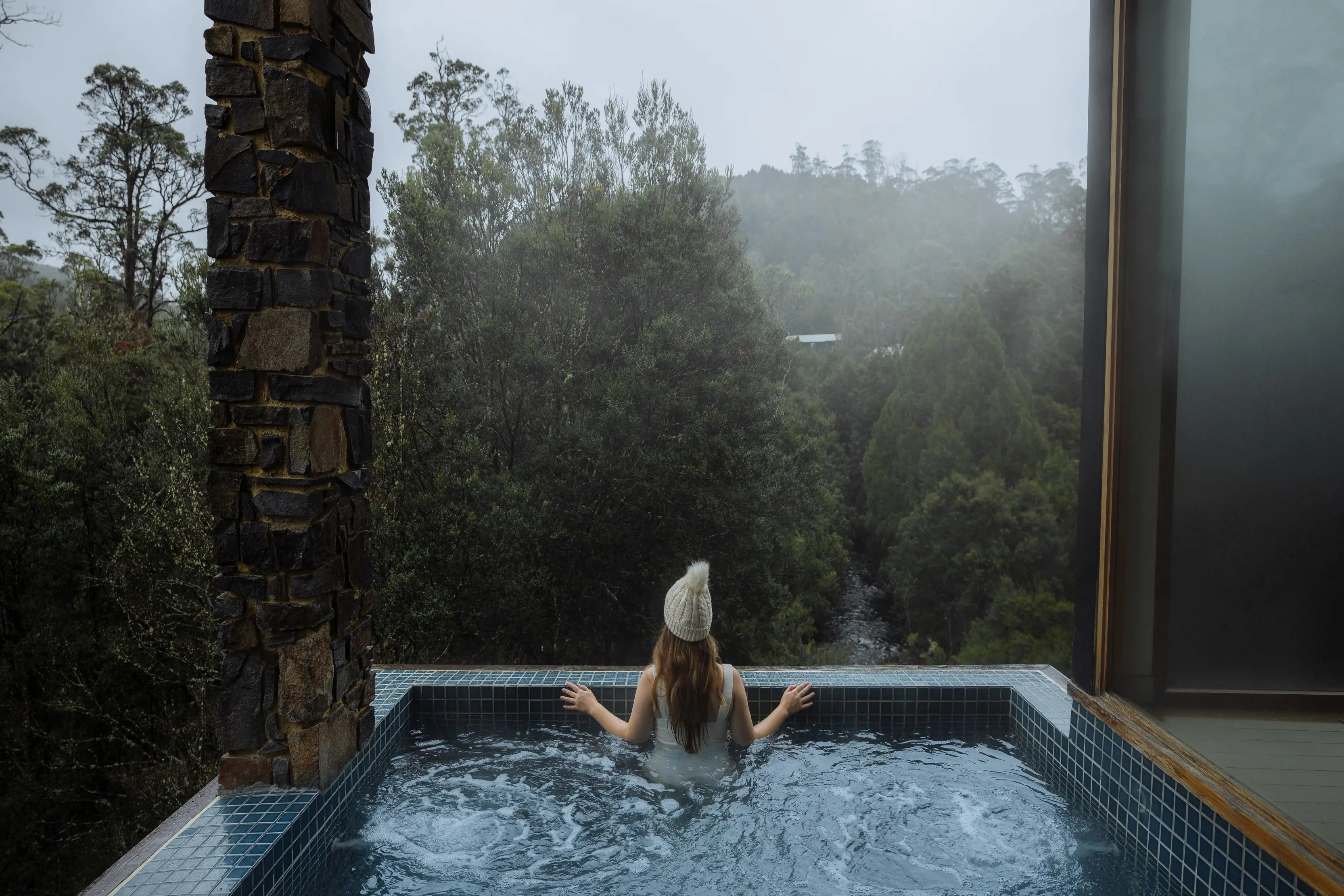 A lady sitting in the spa, wearing a beanie, looking out into the landscape at the Waldheim Alpine Spa at Peppers Cradle Mountain Lodge.