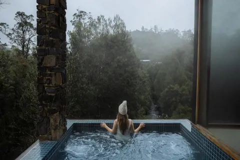 A lady sitting in the spa, wearing a beanie, looking out into the landscape at the Waldheim Alpine Spa at Peppers Cradle Mountain Lodge.