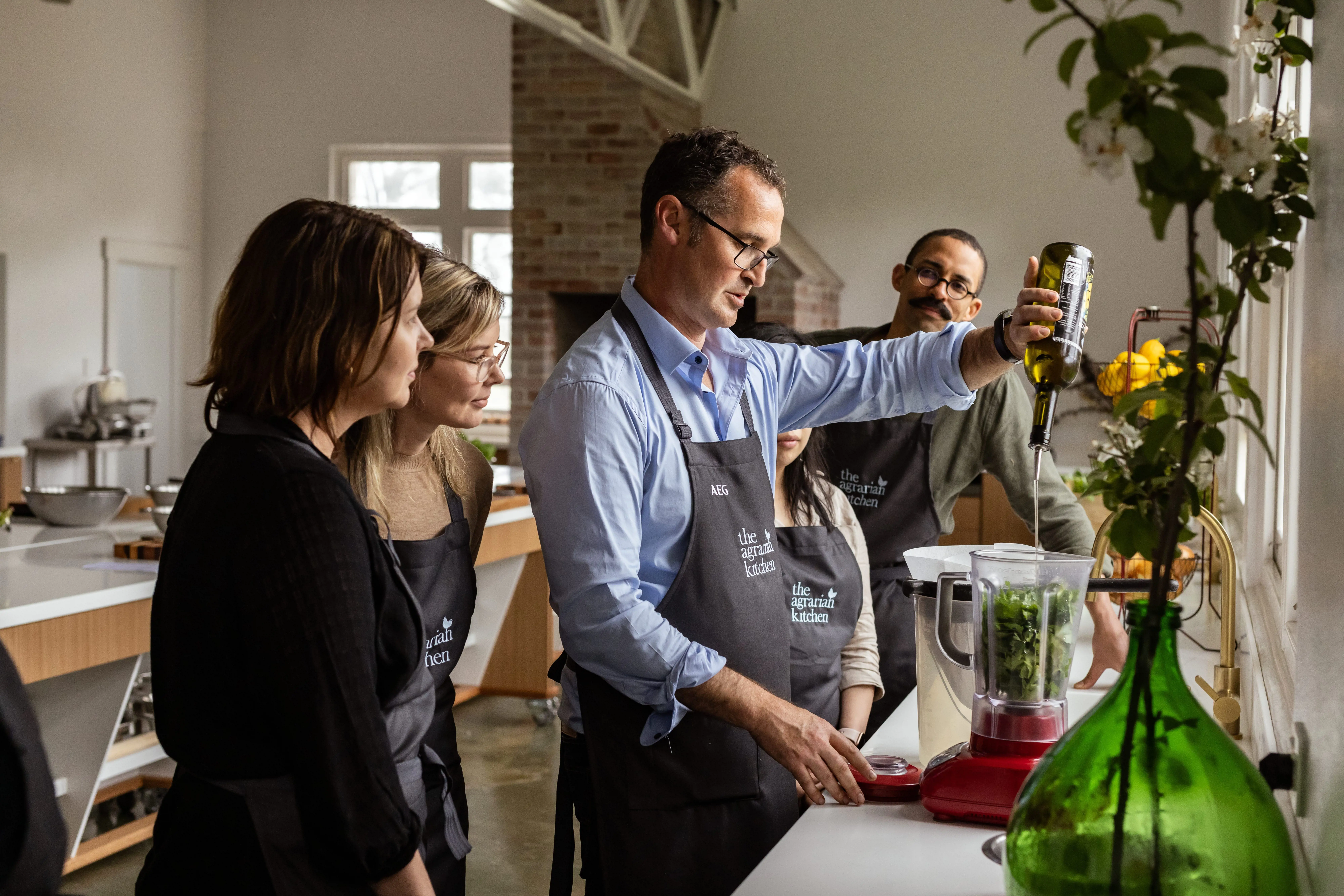 A professional chef pours olive oil into a food processor while a group of students watch over.
