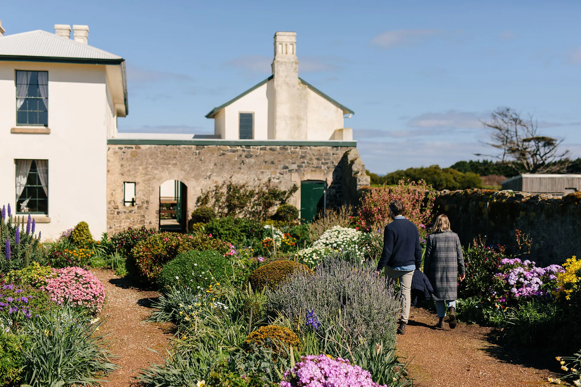 Two people walk through a garden of wildflowers, with red dirt paths backing onto a historic cottage.