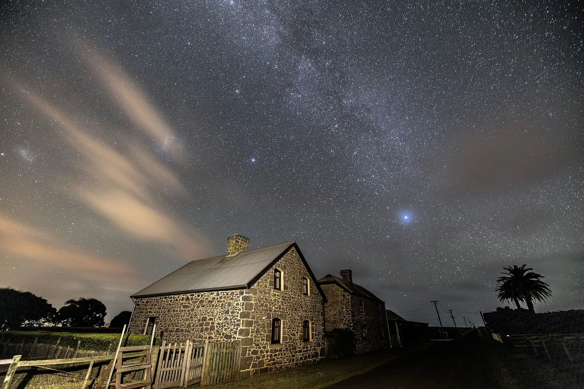 An evocative image captures the Milky Way and other stars above a lit-up heritage stone cottage at night.