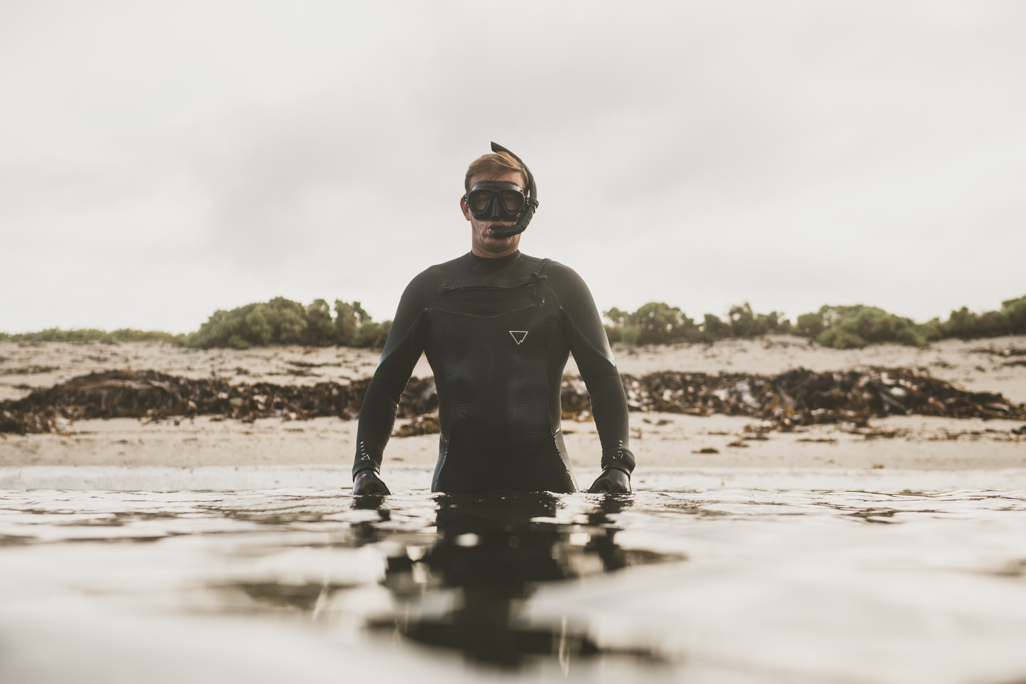 Person in the centre of image, standing in waist deep water, staring at the camera in a wetsuit and a snorkel and goggles on his face.