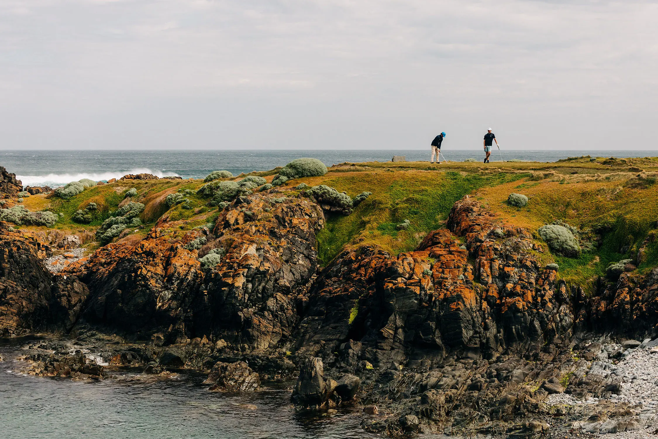 Two golfers stand on a rocky, shrub-dotted outcropping of land, framed either side by the ocean's crashing waves.