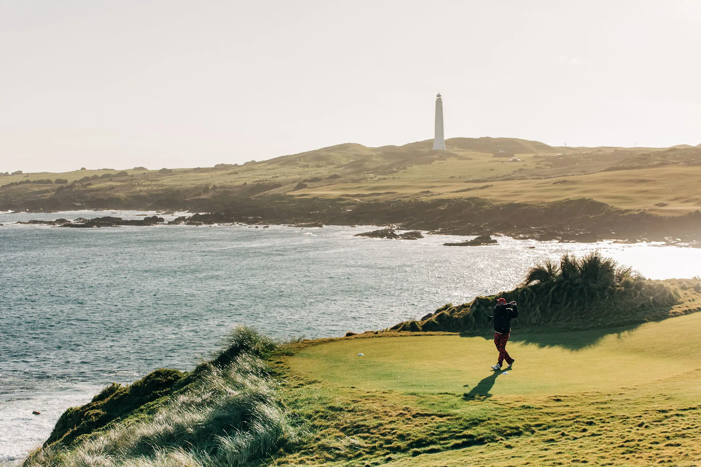 A golfer swings on a green laid out on a small spit of land, looking towards the coast across the water. In the distance a lighthouse rises from the cliffside.