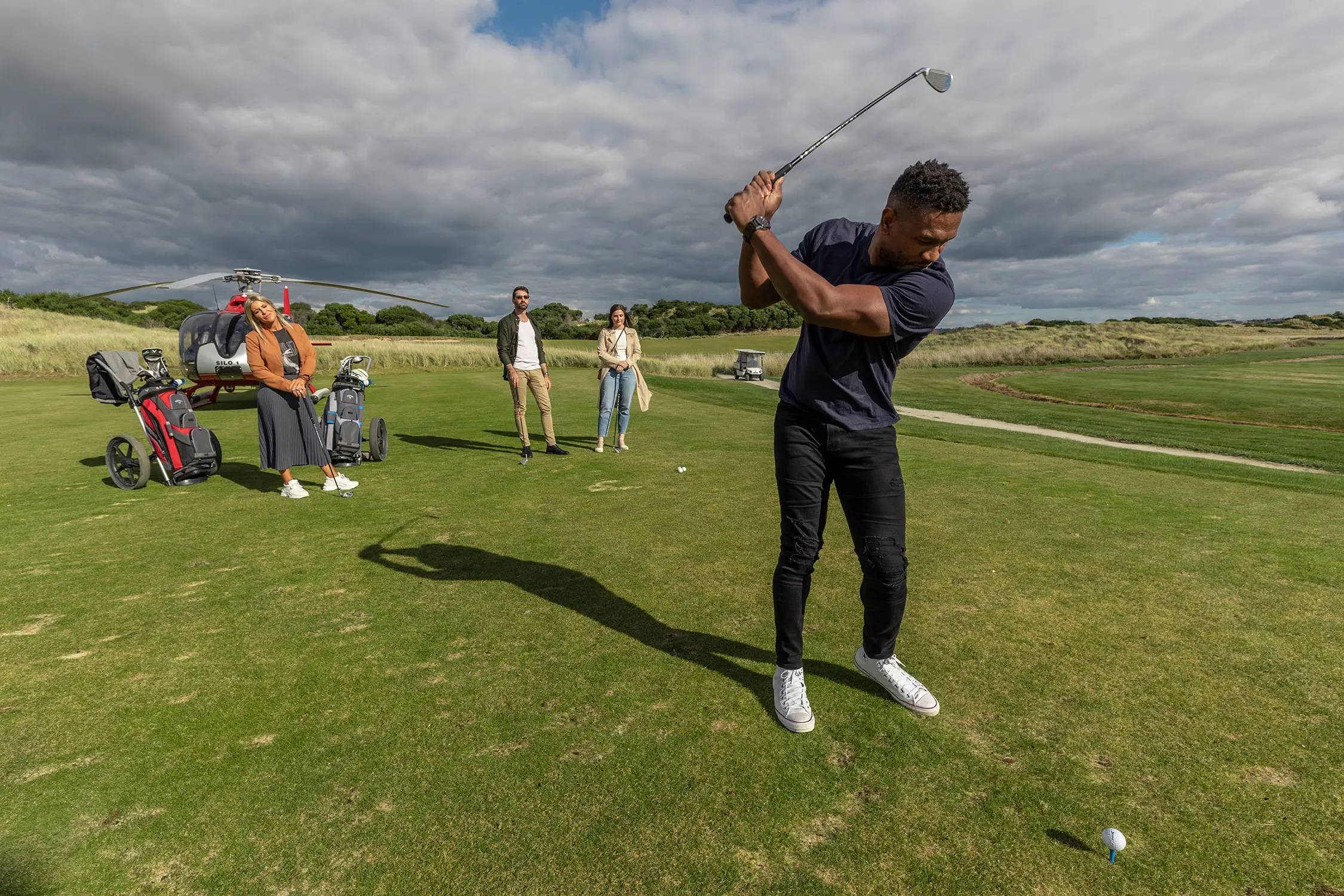 A man swings a golf club, preparing to hit a ball. On the green behind him stand some more people, golf carts and a helicopter awaiting takeoff.