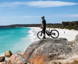A mountain bike rider stands on the orange coloured boulders of the Bay Of Fires, looking out to sea.