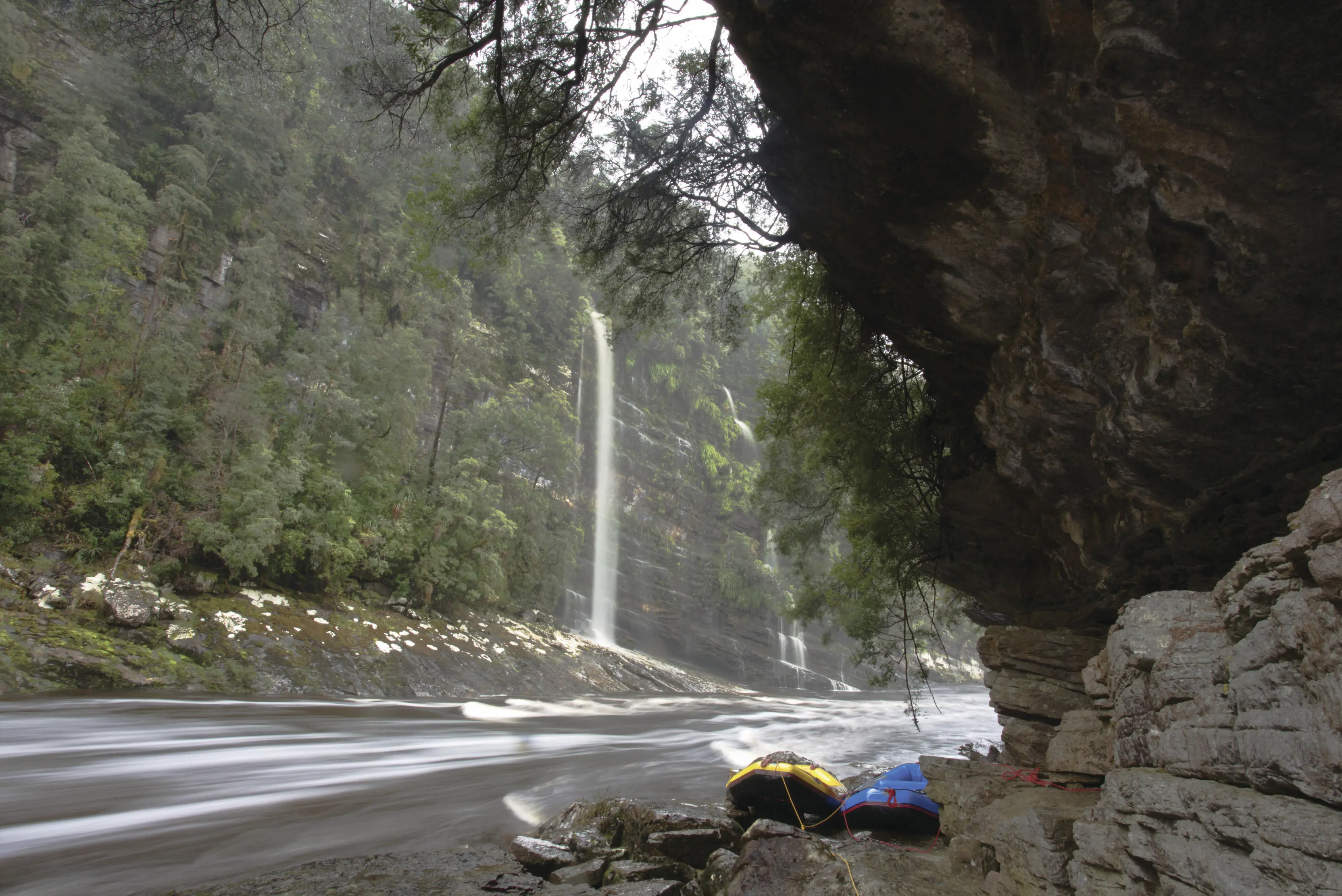 Wide angle image of a blue and yellow raft docked in an area of the Franklin River, with a waterfall in the background.