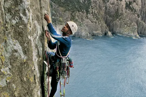 Jaw Dropping image of a person rock climbing up Totem Pole, Cape Hauy. A stunning landscape of mountains and ocean surround them.