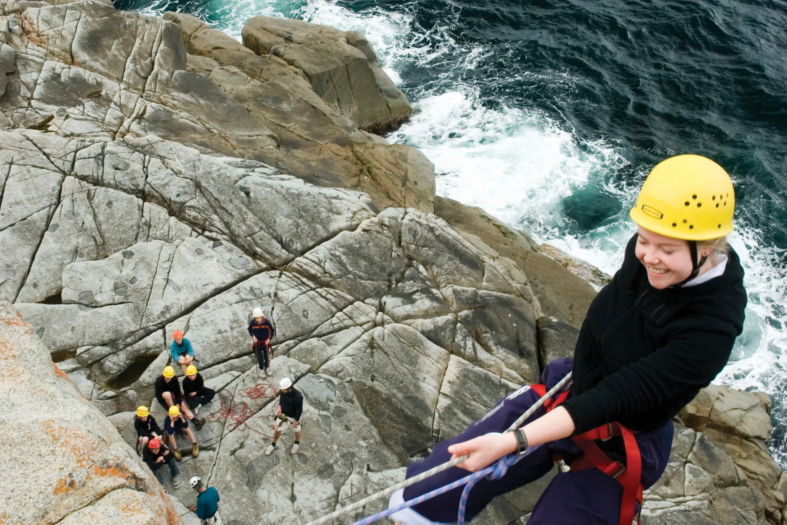 A woman wearing safety equipment and a harness abseils down a rocky cliff near the ocean. A group watch from below.