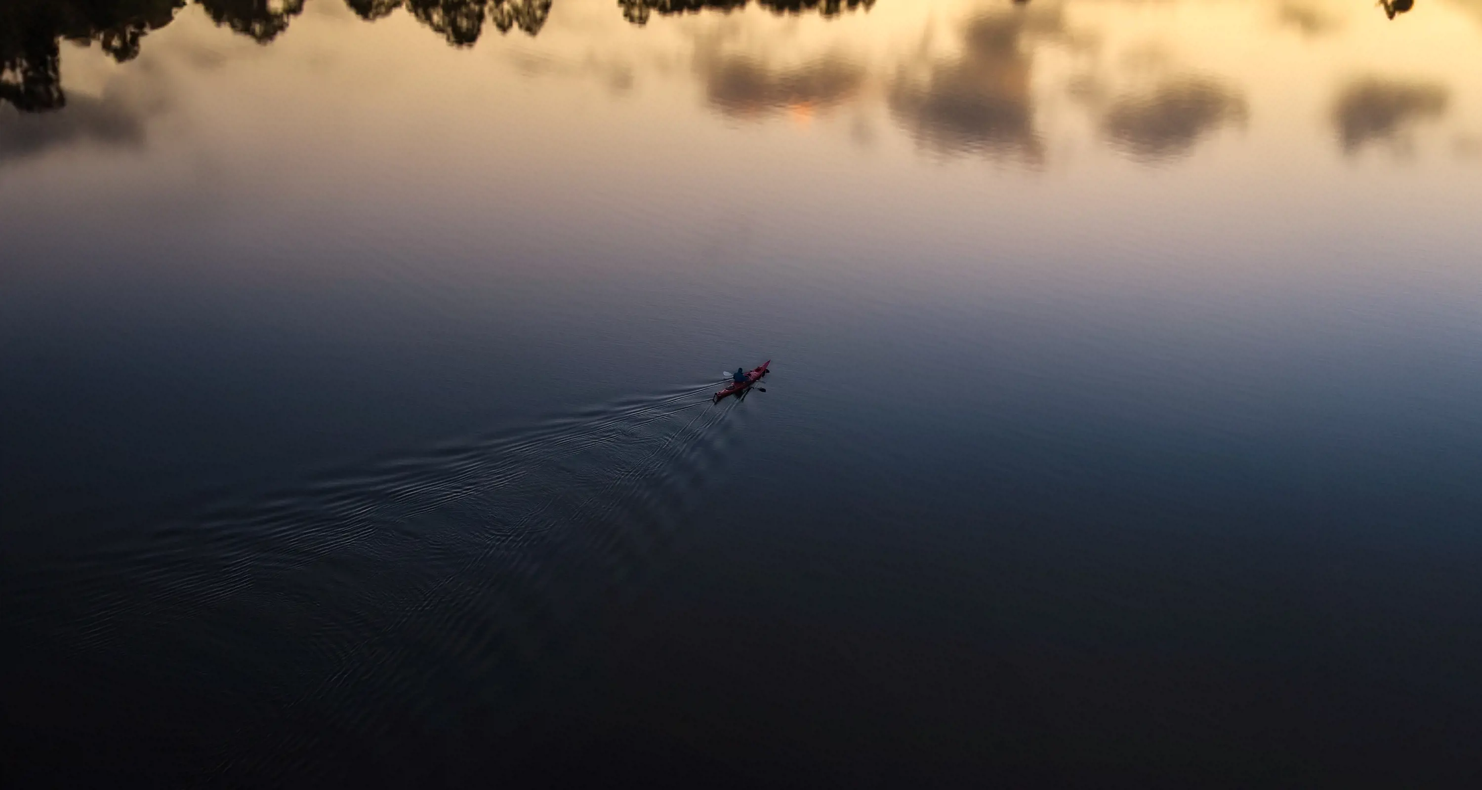 A kayaker floats out into the middle of a wide river, with the reflections of trees in the water. 