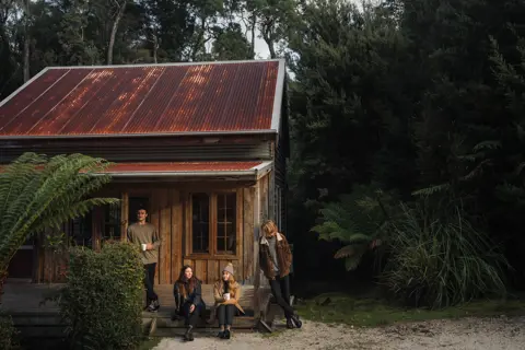 Group of four sit outside one of the Corinna huts, amongst dense bushland.