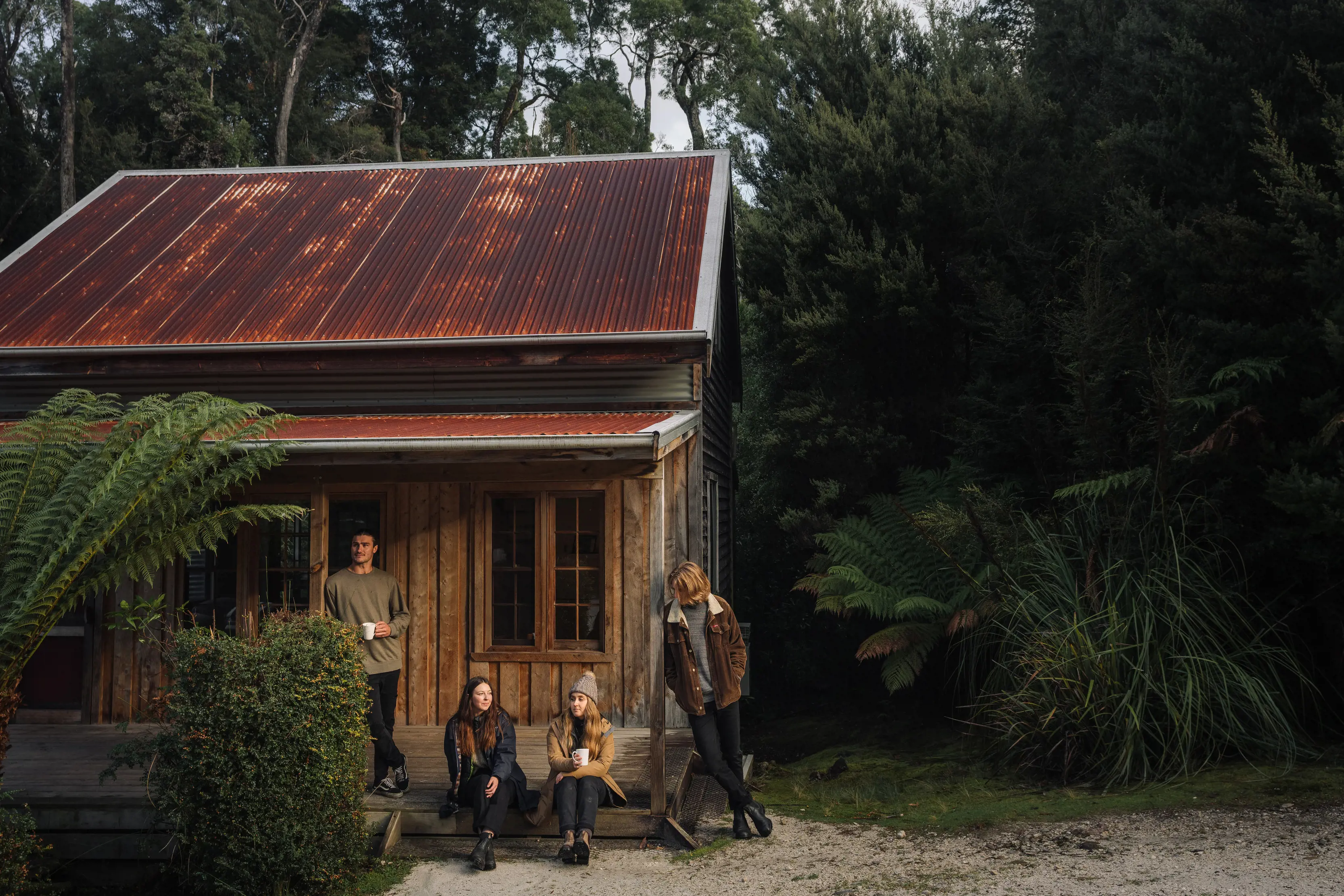 Group of four sit outside one of the Corinna huts, amongst dense bushland.