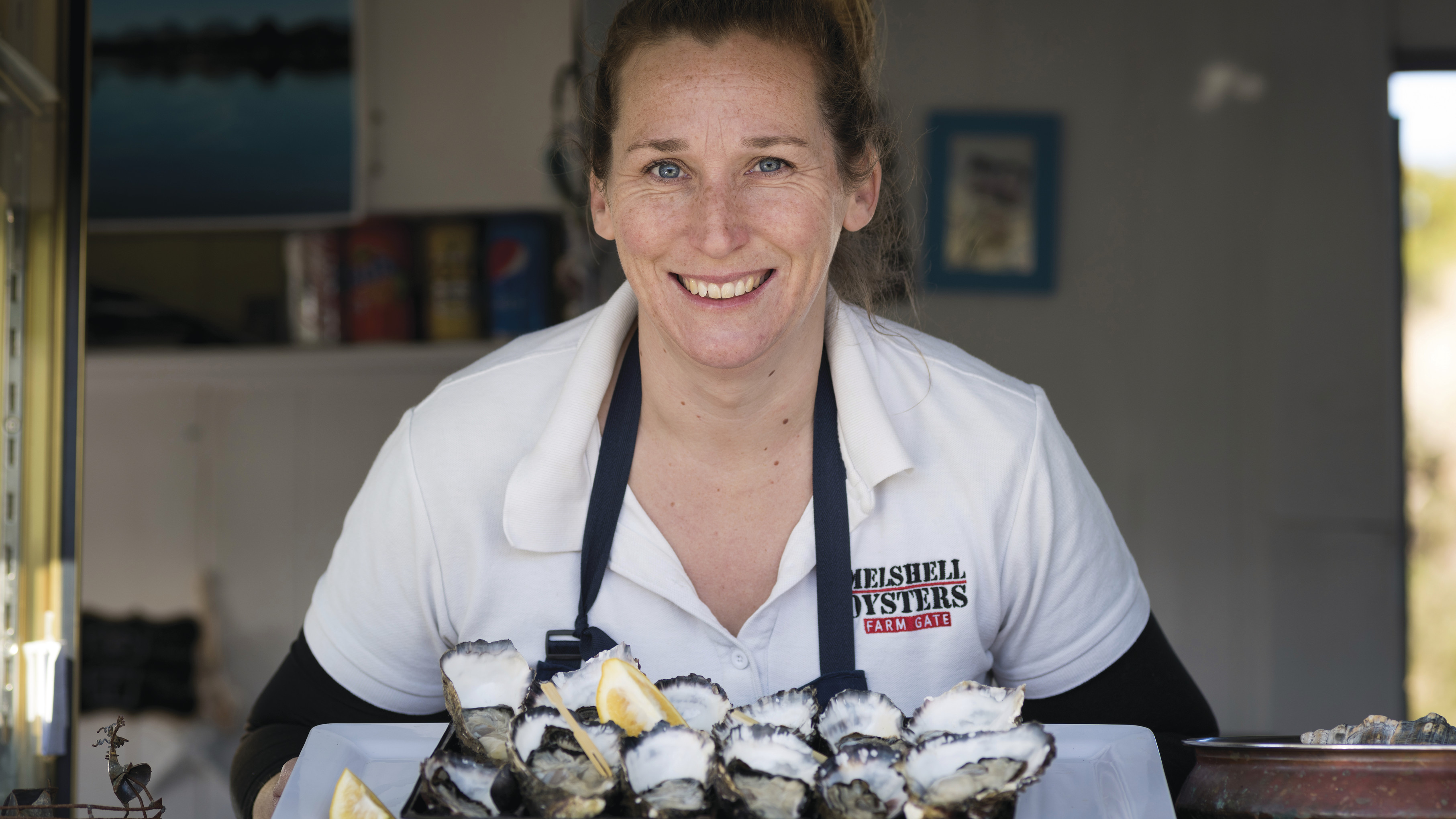 Cassy Melrose smiling at the camera and holding a large platter of fresh oysters with lemon wedges.