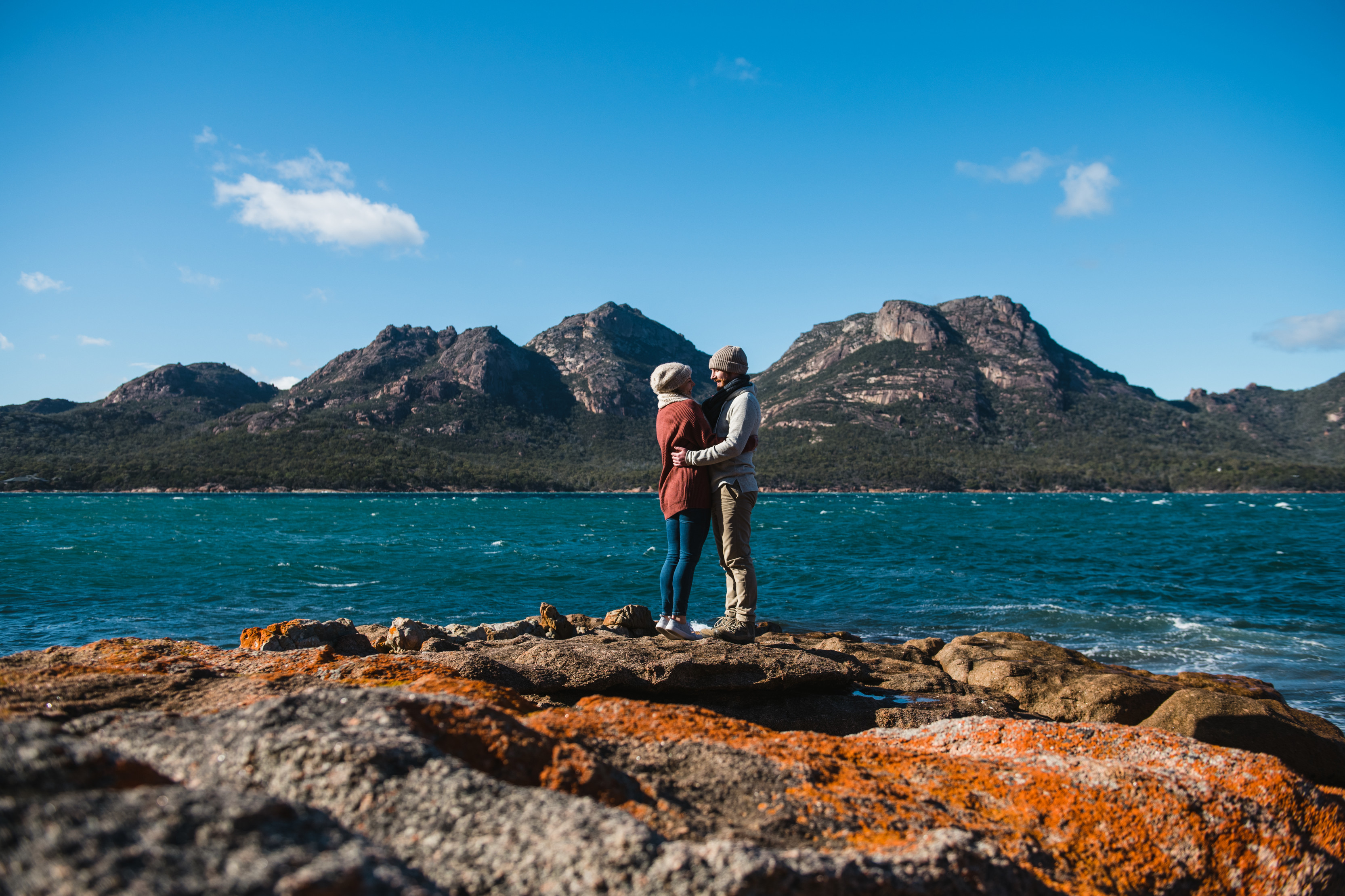 Couple hugging each other on the point at Coles Bay, overlooking the calm, crystal clear water of Oyster Bay.