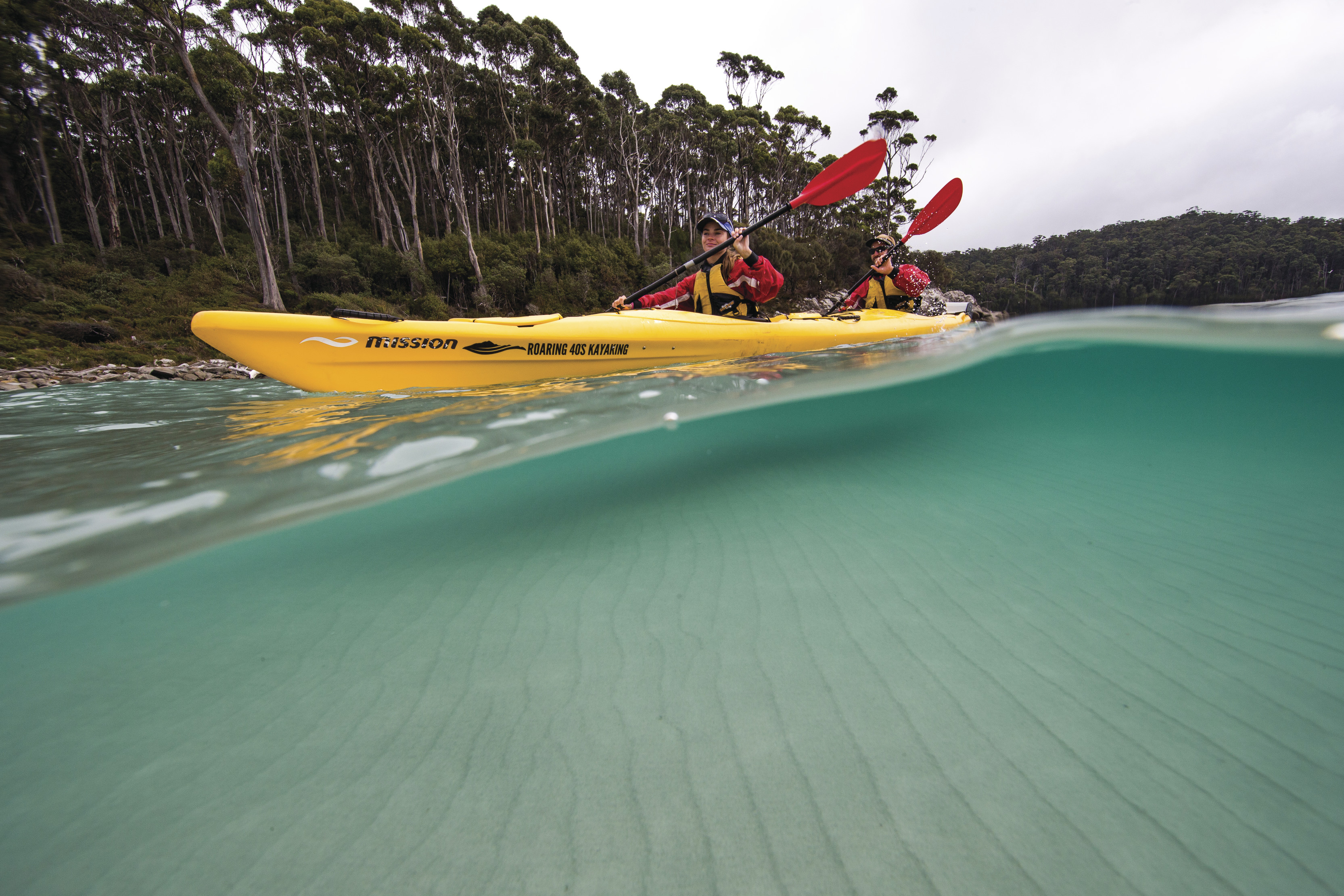 Stunning wide angle, over/under image of two people in a twin kayak, paddling along pristine, blue water.