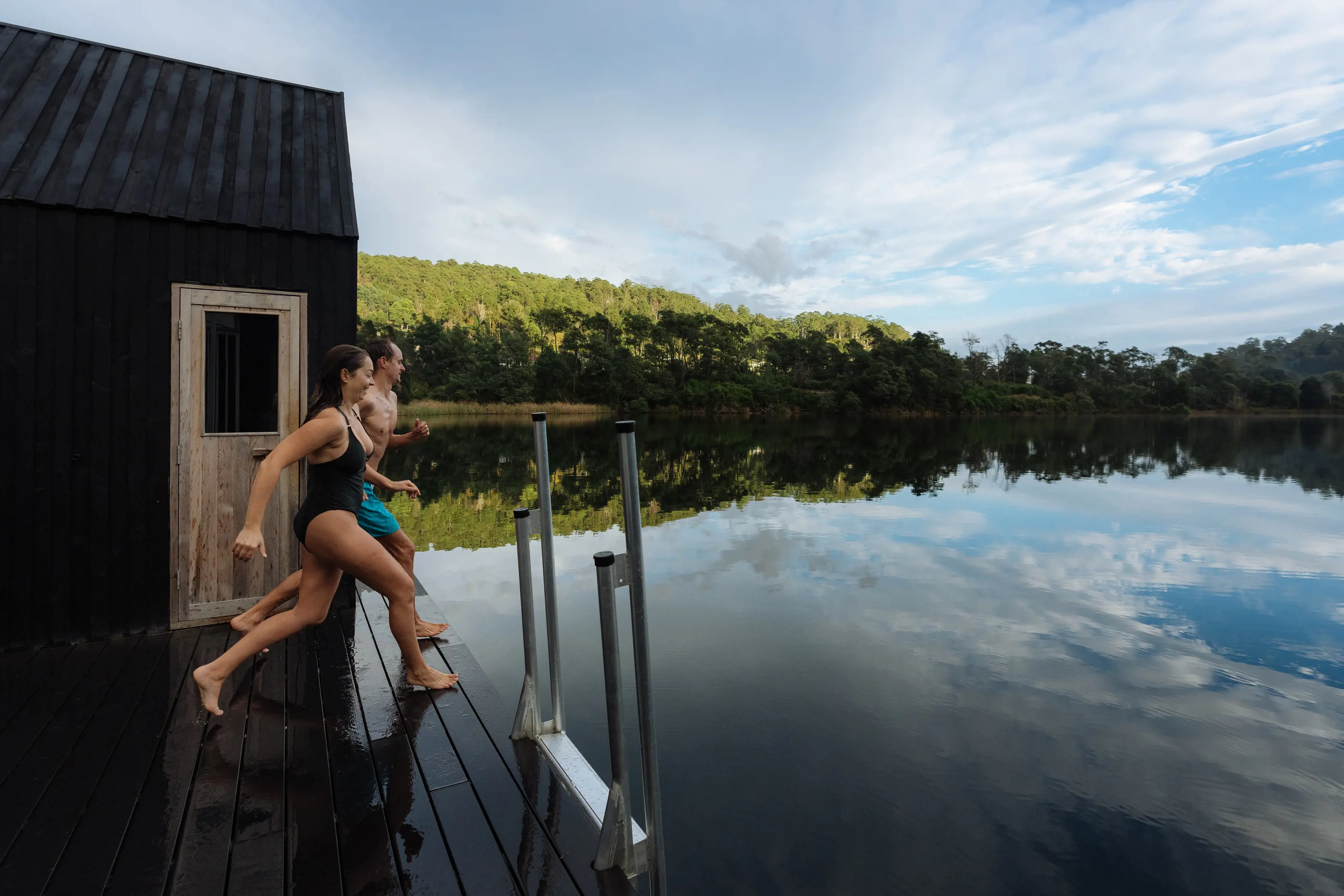 Couple running along a pier, about to jump into the Floating Sauna Lake Derby, surrounded by stunning wilderness.