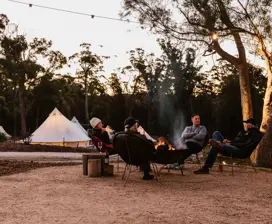 Four people sitting around a camp fire at Bay of Fires Bush Retreat. Pitched, white tents in the background
