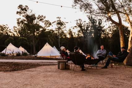 Four people sitting around a camp fire at Bay of Fires Bush Retreat. Pitched, white tents in the background