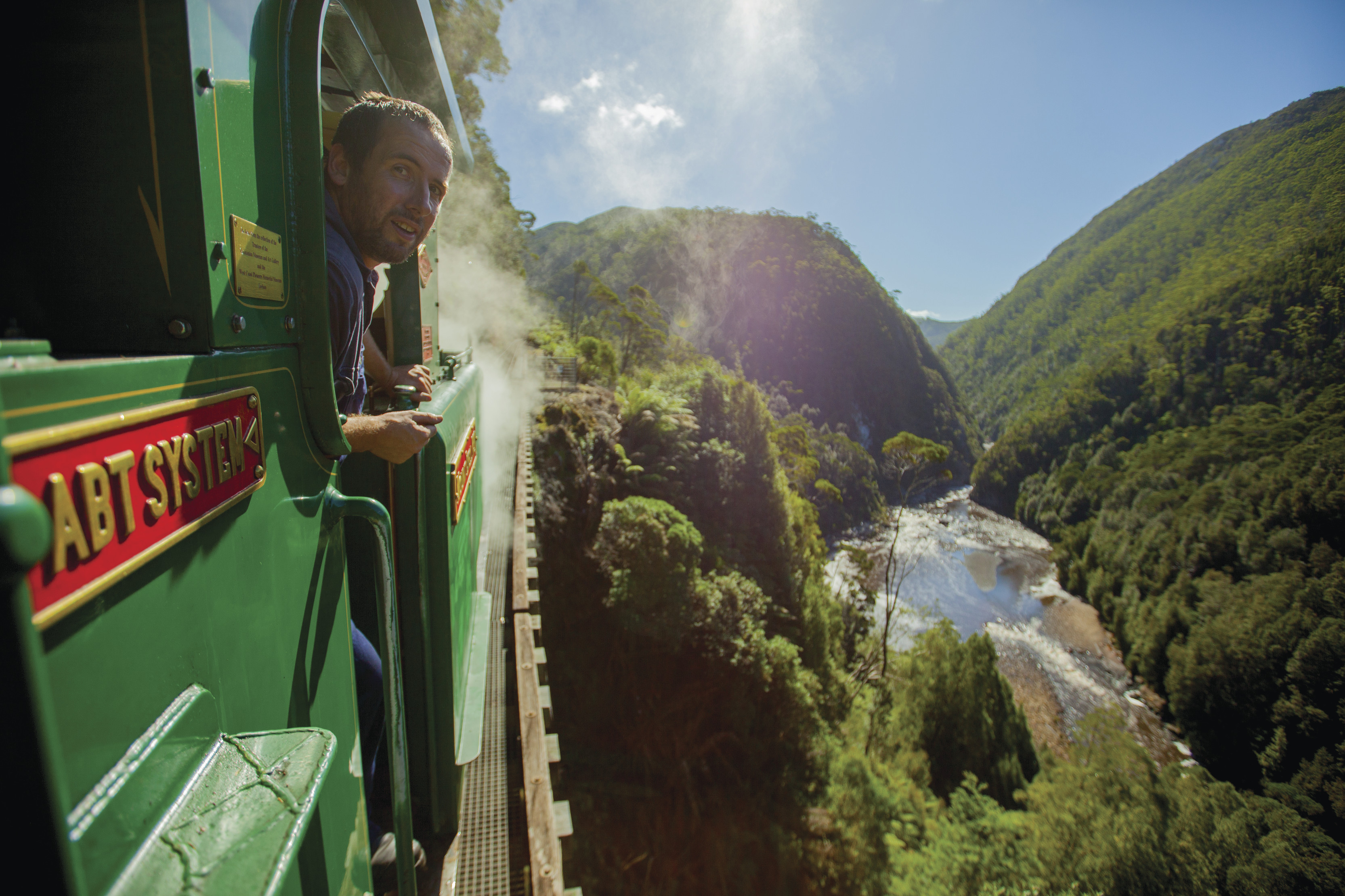 "Person sticking their head out of the train, back towards the camera, surrounded by lush green forest and water, with West Coast Wilderness Railway. "