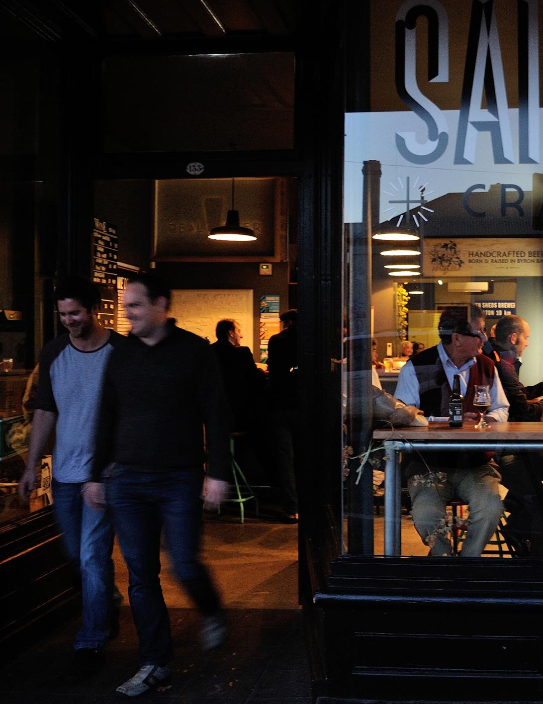 Two young men leave the entrance of a craft beer bar. An elderly man is sitting in the window at a high bench, talking with other customers.