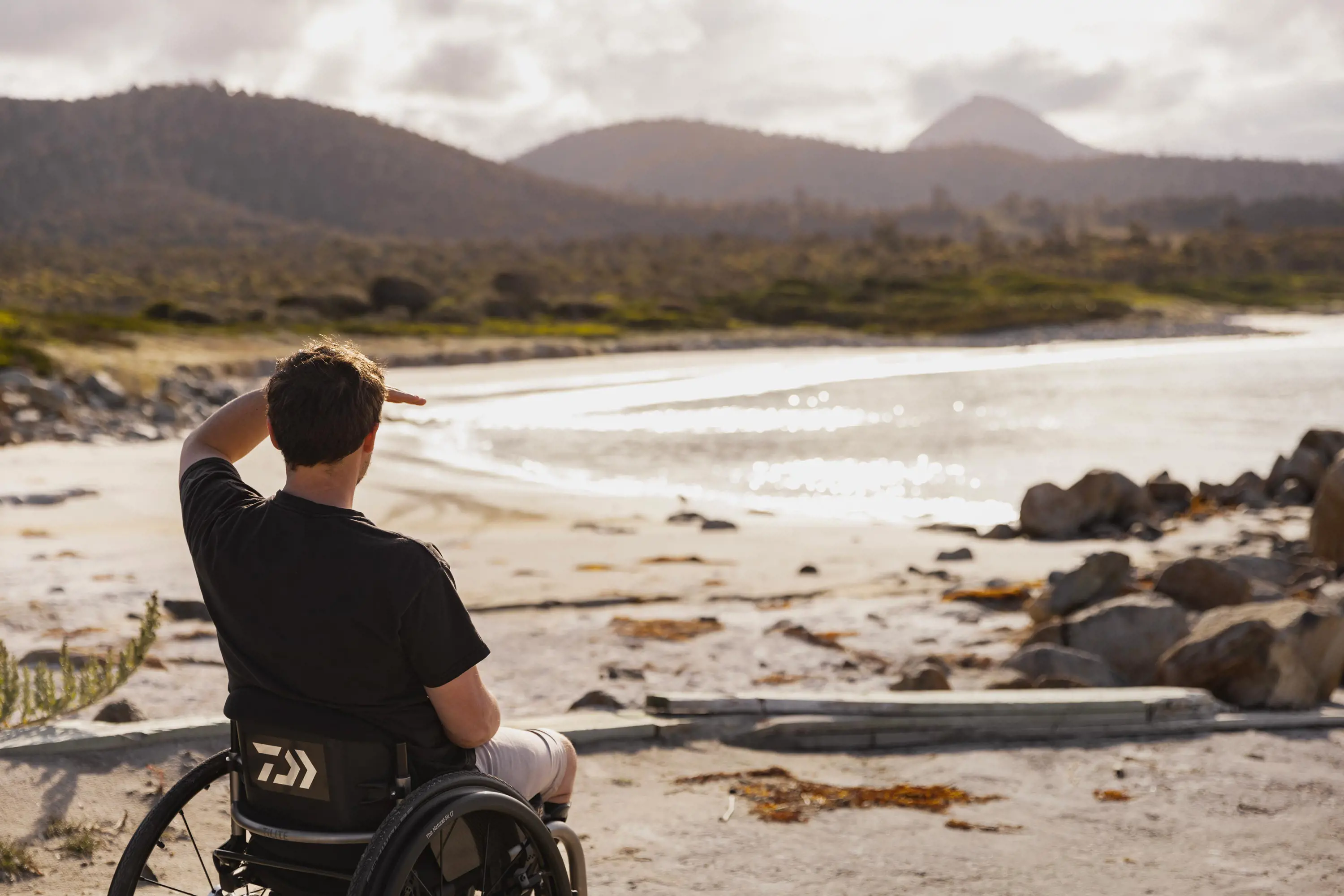 A young man wearing a t-shirt and sitting in a wheelchair looks out over a beach on a sunny day.
