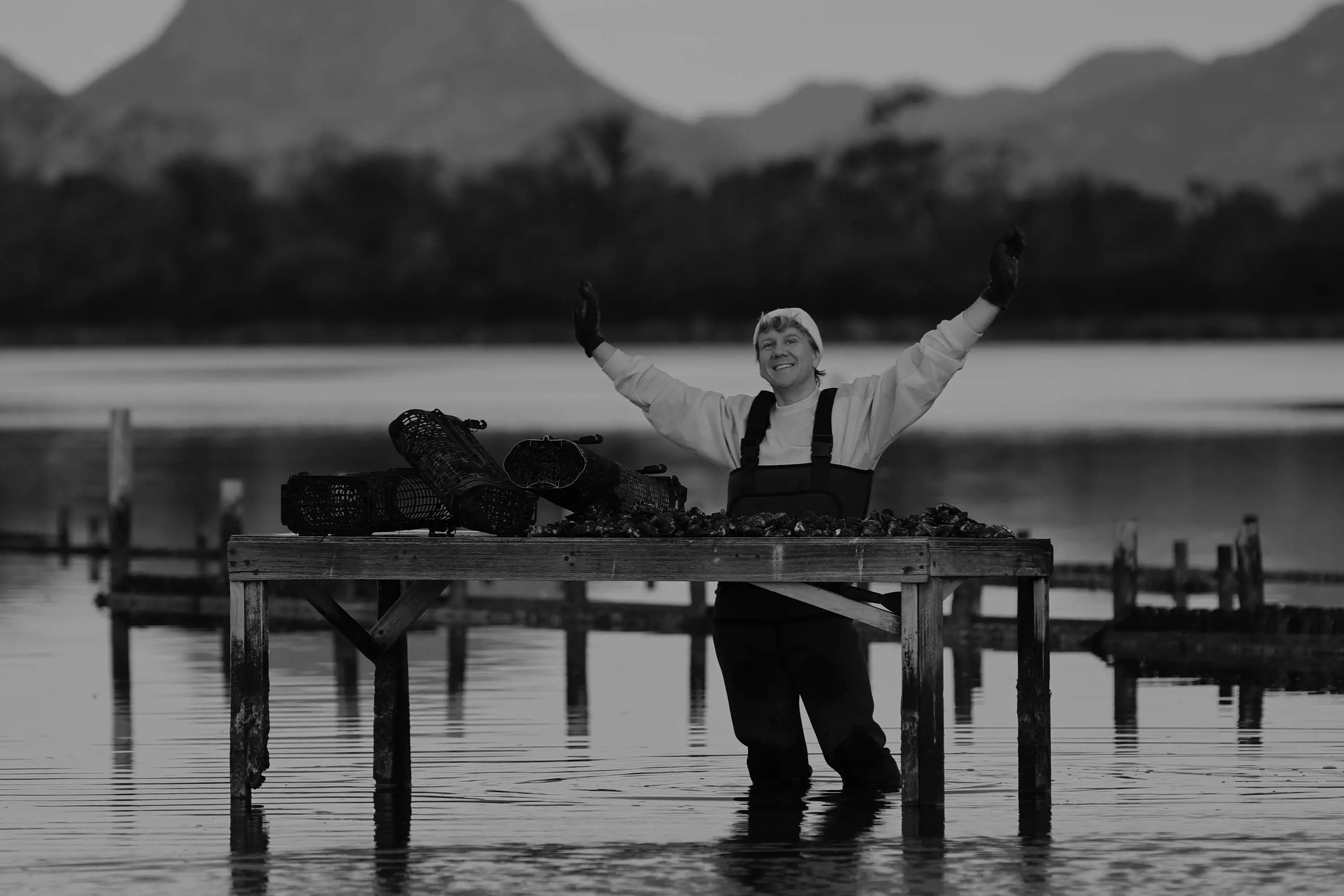 A man wearing waders stands in knee-high water next to a table with oyster nets.