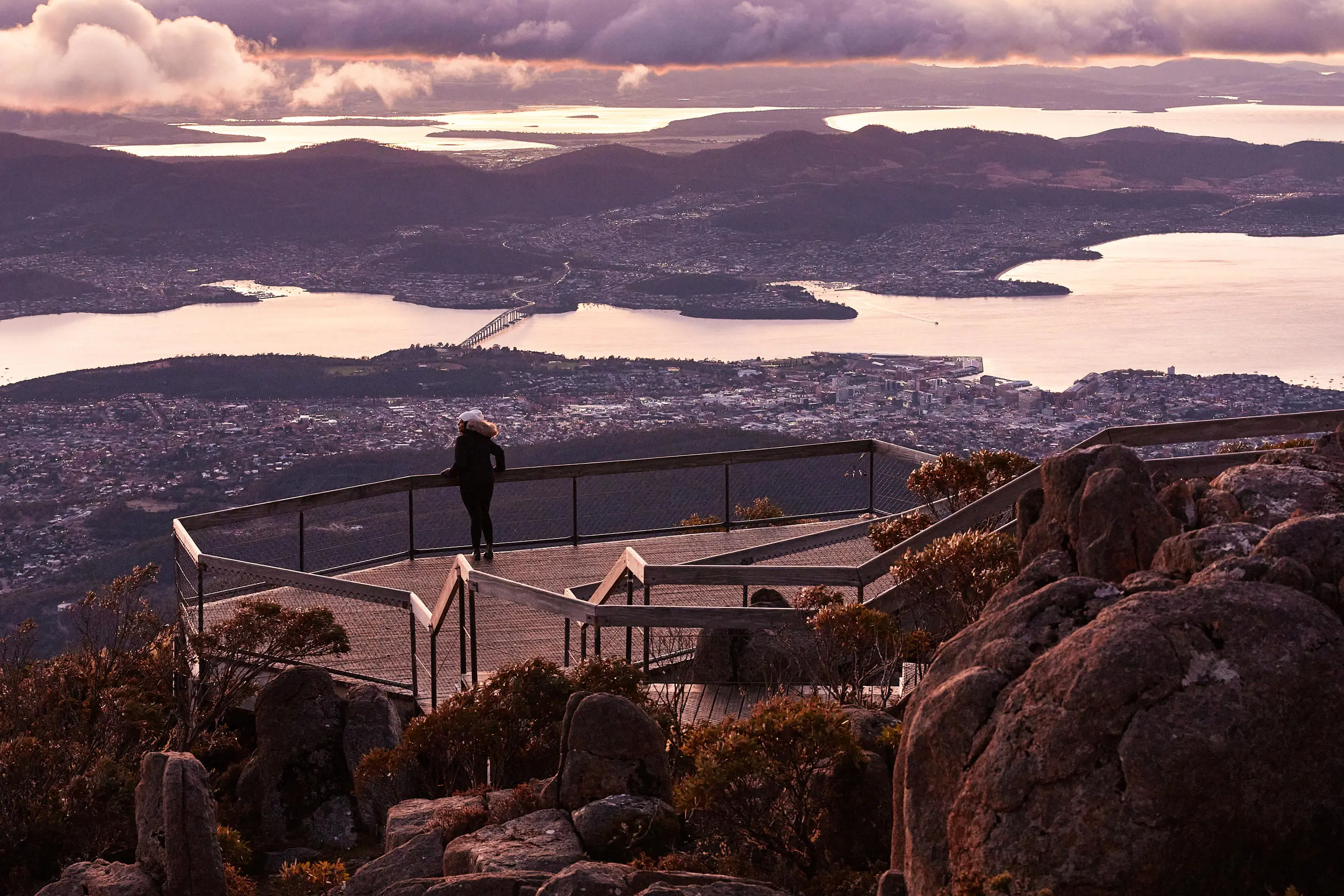 A young woman stands on a viewing platform and looks down on Hobart city and the River Derwent at dawn.