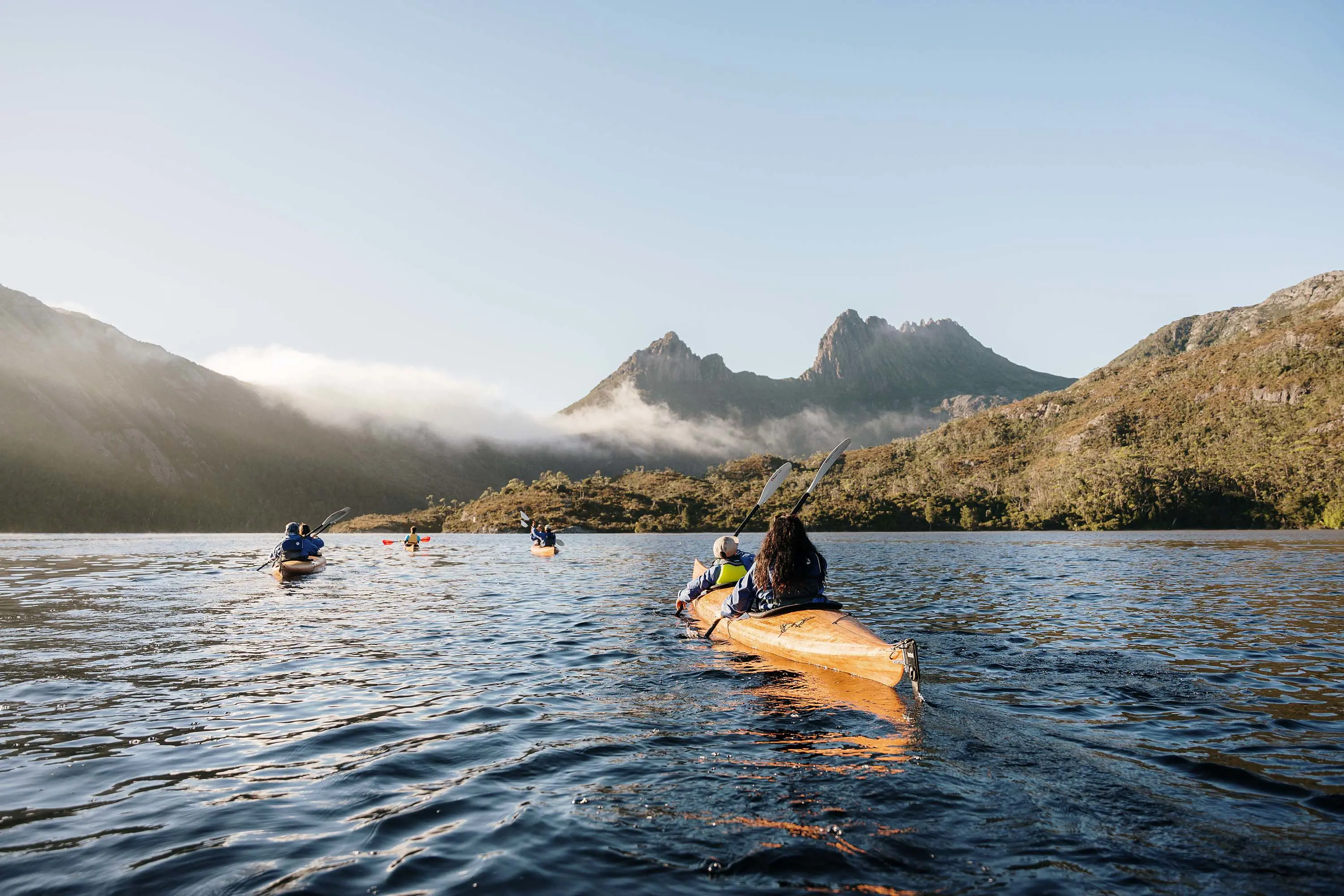 Four canoes float across a lake in front of the tall, dramatic peaks of Cradle Mountain on a clear day.