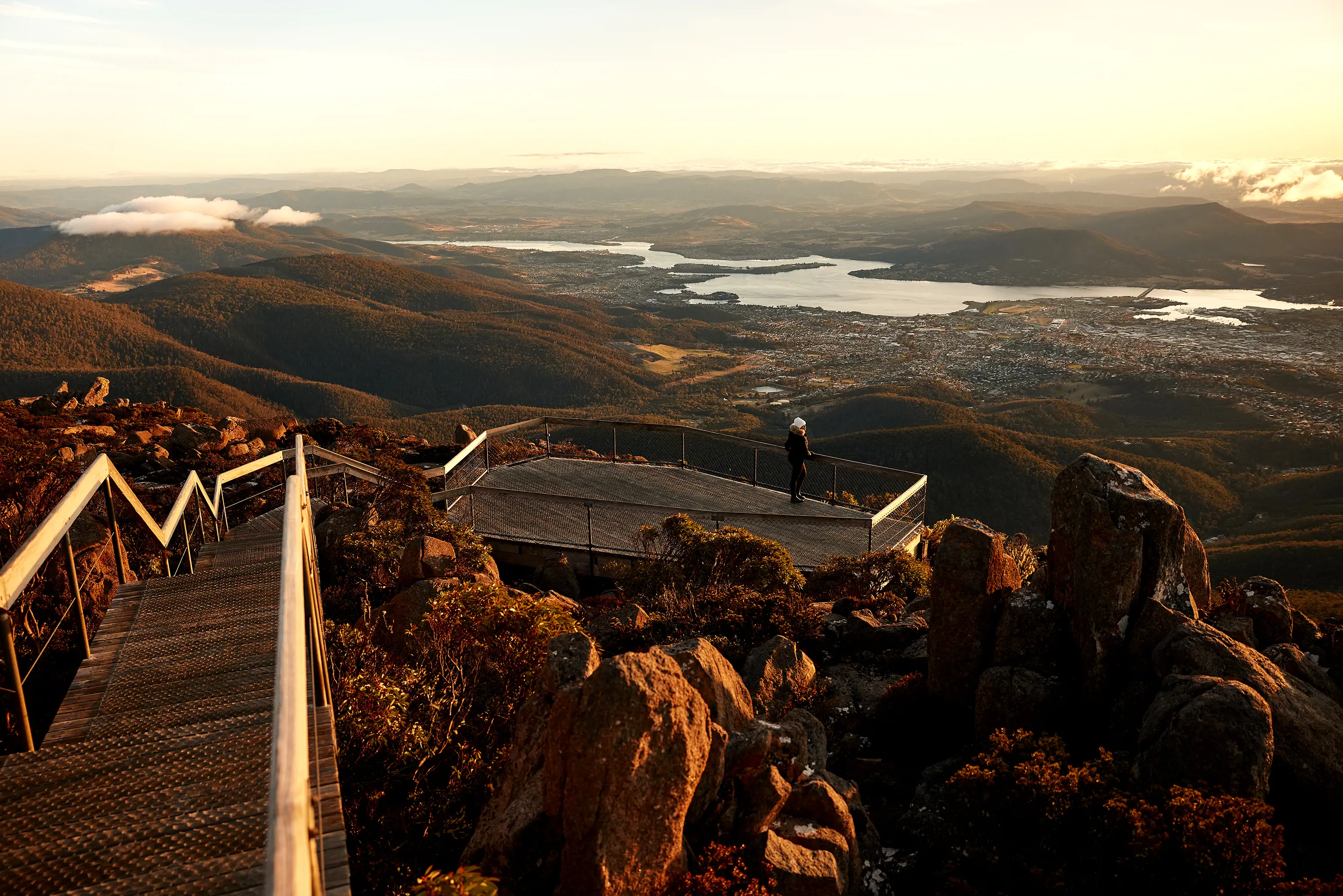 A woman stands at the end of a viewing platform on the side of a mountain overlooking Hobart and the River Derwent at sunrise.